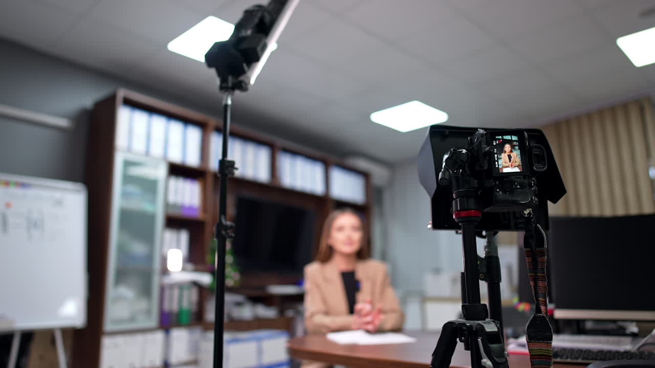 Day-light lamp on the stand and camera on tripod are set in the office. Woman in beige jacket recording blog content. Blurred backdrop.