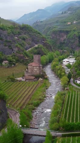 vertical drone ascends alongside castel roncolo, showcasing its medieval stone towers, crenellated walls, and ivy-covered facades perched atop a rocky spur above the talvera stream.