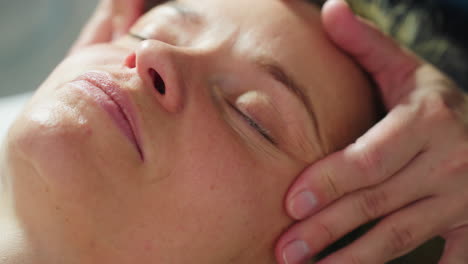 close up of recipient lying face up on massage bed as female skin therapist gently massages facial contours around eyes cheeks and forehead under soft spa lighting for relaxing rejuvenation session