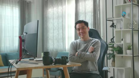 asian cameraman in long sleeved t-shirt and black pants crossing his arms and smiling to camera while working with desktop computer at home.