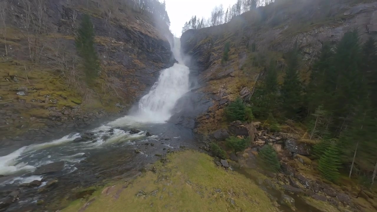 FPV-drone suurella nopeudella alas vesiputousta norjalaisessa metsässä, paljastaen putoavan veden virtauksen ja joen.