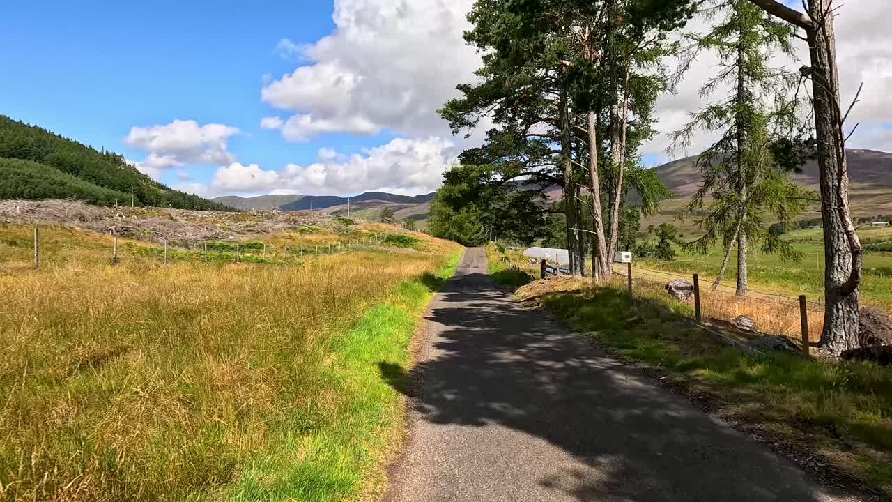 A vehicle travels along a narrow country road through scenic Clova, Angus, Scotland, on a bright summer day with clear skies and rolling hills