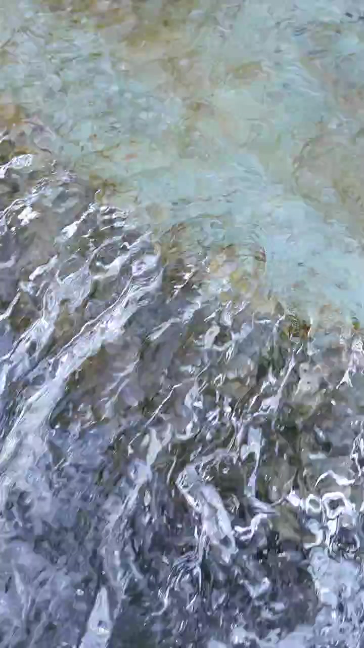Vista ravvicinata di acqua trasparente che scorre su rocce texturizzate in un ambiente naturale all'aperto, con telecamera stabile e luce diurna soffusa che crea un'atmosfera tranquilla