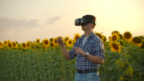 a young farmer in plaid shirt and jeans uses vr glasses on the field with sunflowers for 3d moderling. these are modern technologies in summer evening.