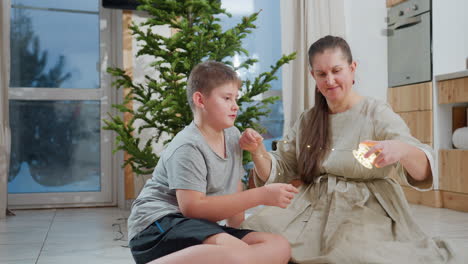 mom happily rolls christmas light and places it around son's neck as they both smile, enjoying holiday moments together. family bonding in front of christmas tree with festive atmosphere