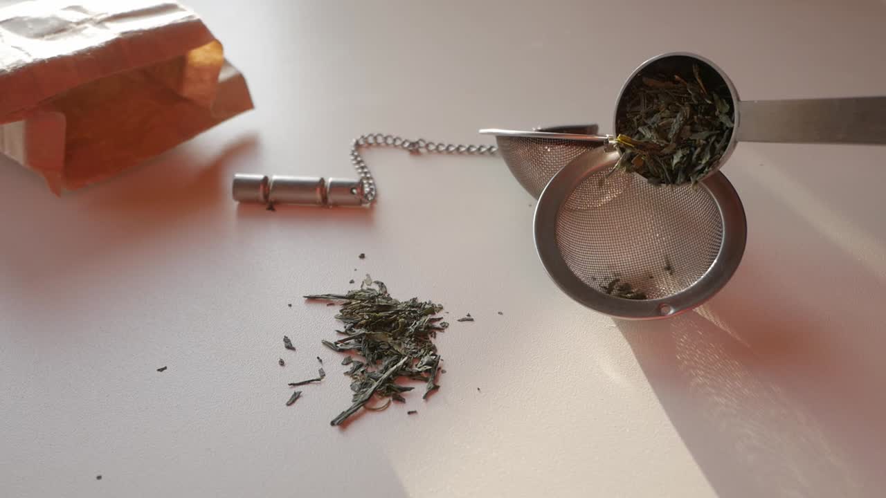 Fresh green tea leaves being prepared for brewing with a tea infuser and a paper bag