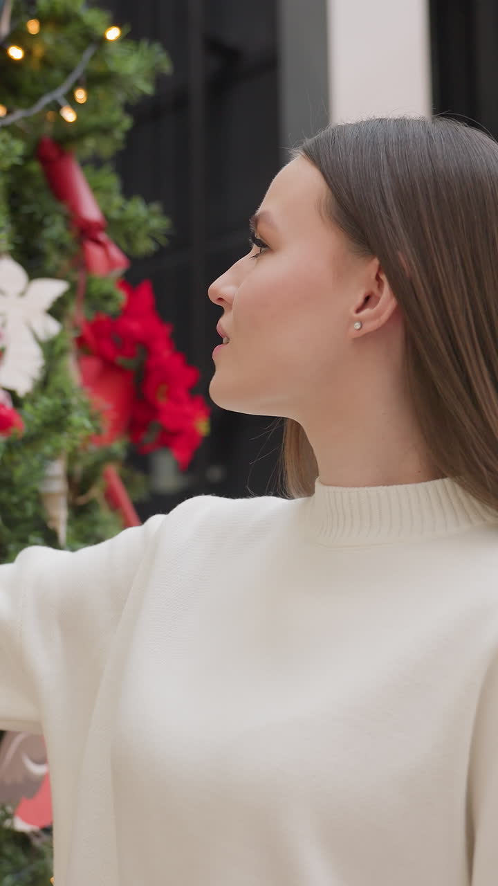 Young woman walking towards festive Christmas tree, holding green ornament with care while admiring decorations in the background of shopping mall with soft lighting