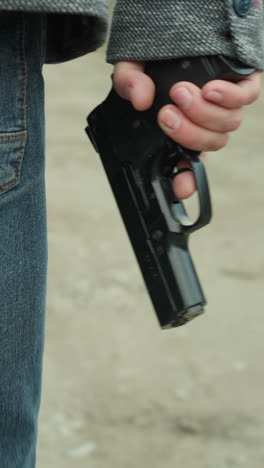 vista cercana de una persona en vaqueros azules y una chaqueta gris sosteniendo una pistola mientras camina por un camino de tierra