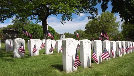 Vista-Lateral-Del-Día-Conmemorativo-De-Las-Lápidas-De-Los-Veteranos-De-La-Guerra-Civil-En-Un-Cementerio-Mientras-Las-Banderas-Americanas-Soplan-En-El-Viento-Bajo-Un-Cielo-Azul-Nublado-Y-árboles-Verdes