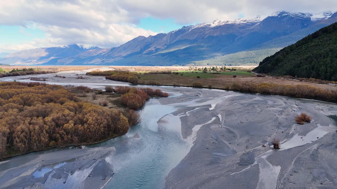 Drone glides above braided river, autumn trees, and snow-capped mountains under soft daylight