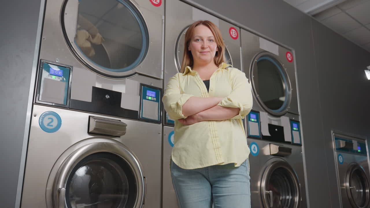 Supervisor posing with hands folded, while standing before industrial cloth dryers, clothes rolling behind glass, stainless machines with glowing controls, modern laundromat scene showcasing service