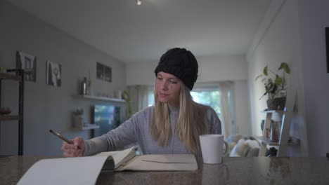 a beautiful blonde woman writing on a notebook and drinking a hot beverage at home