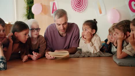 A-man-with-gray-hair-in-a-purple-T-shirt,-a-teacher-reads-a-book-to-his-students-who-lie-on-the-floor-on-special-pillows-and-listen-carefully-to-their-teacher-in-a-club-for-preparing-children-for-school