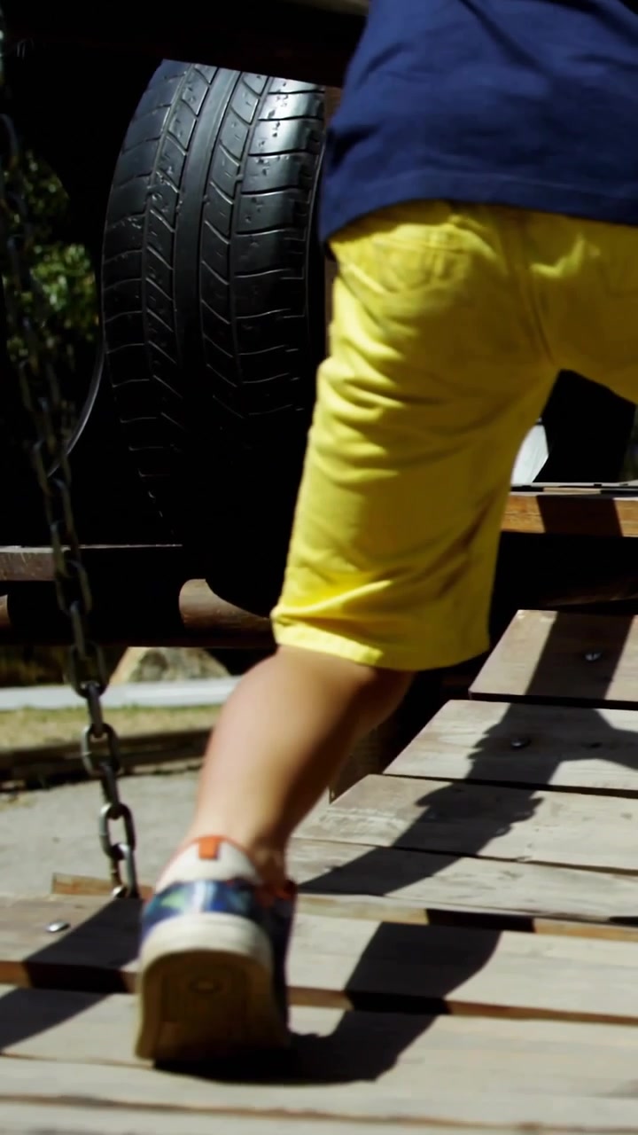 Vertical video: Boy yellow shorts stepping plank bridge, chain swinging, safety cues aiding play