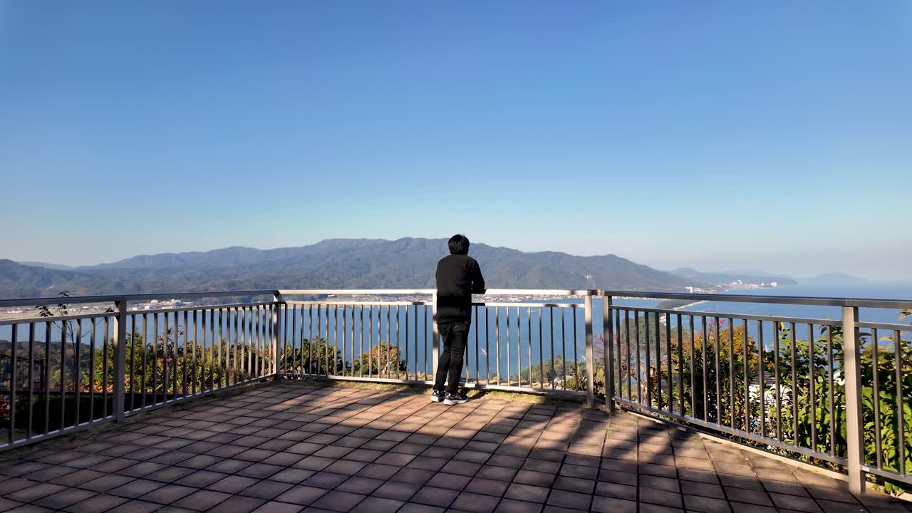 Young man enjoying panoramic view of Amanohashidate sandbar from a hilltop viewpoint, appreciating the beauty of the Japanese landscape