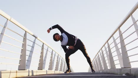 african american man exercising outdoors, standing on footbridge stretching from waist