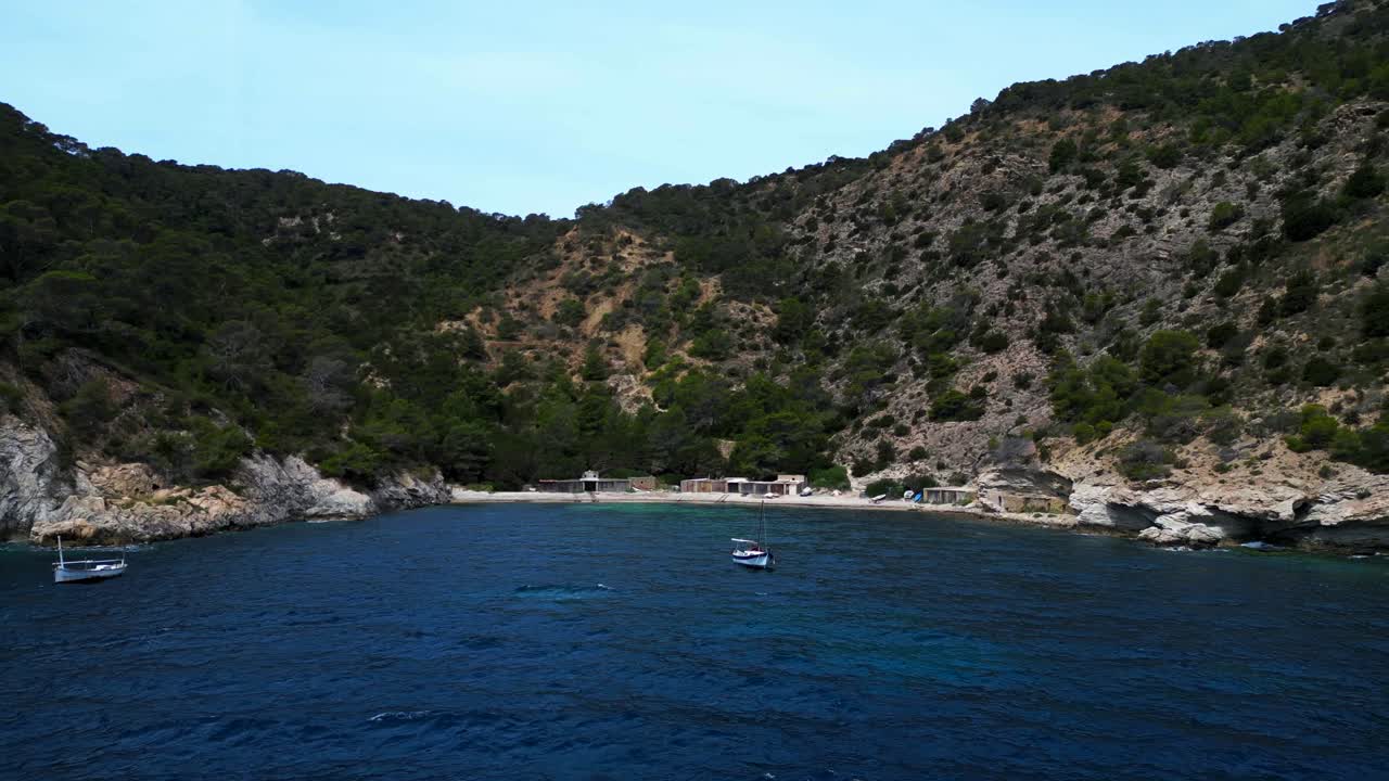 Sailboat floating on calm blue water near a hidden beach bay with some fishermen's houses in Ibiza. Magic aerial view flight fly reverse panorama overview drone