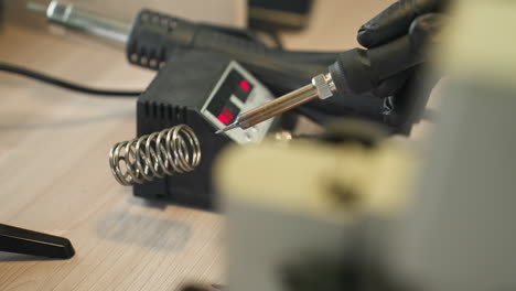 a close-up view of a gloved hand holding a soldering iron, placed in a holder next to a powered soldering station, and cleaning the soldering mouth