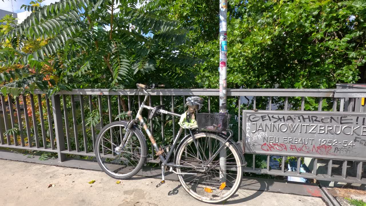 A single bicycle remains parked against a graffiti-covered bridge railing in Berlin, with lush green trees and bright daylight. Camera is static