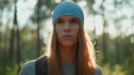 lady in blue headscarf stands in forest, deep in thought, as sunlight creates a radiant glow around her face, she appears calm, with her hair illuminated by the sun filtering through trees