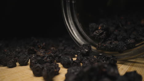 Glass-Jar-of-Dry-Ripe-Wild-Blueberries-Fruit-Lying-Down-on-Wooden-Surface,-Heap-of-Dried-Bilberries-Scattered-on-Table,-Close-Up-Probe-Lens-Studio-Shot,-Panning-Tracking-Sideways-View