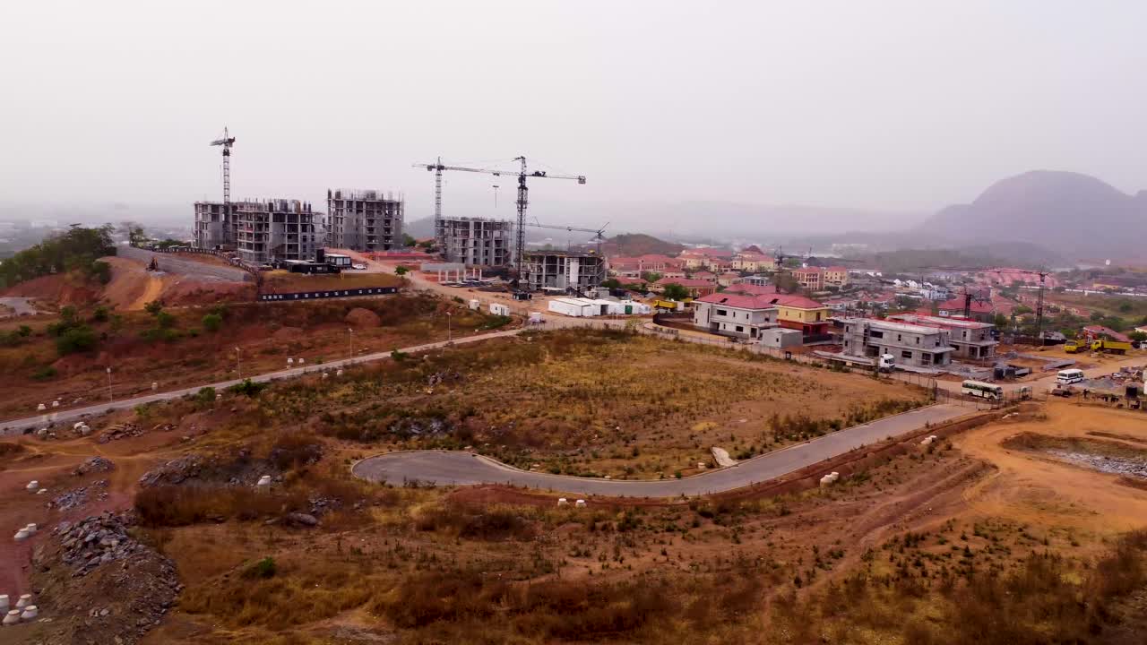 Aerial establishing pan overview of developing urban area in Asokoro, Abuja, showing road networks, buildings, and distant hills with tall construction cranes