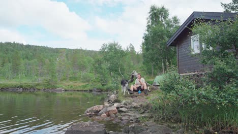 Man-With-His-Alaskan-Malamute-Dog-Sitting-Relaxing-Next-To-The-River-Bank-In-Anderdalen,-Senja,-Norway