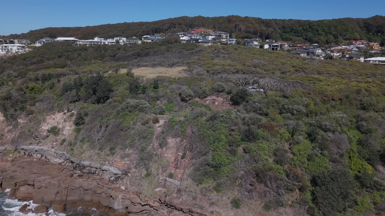 Coastal drone ascend to reveal curved rocky beach and homes on cliff edge, calm water and bright early sunlight
