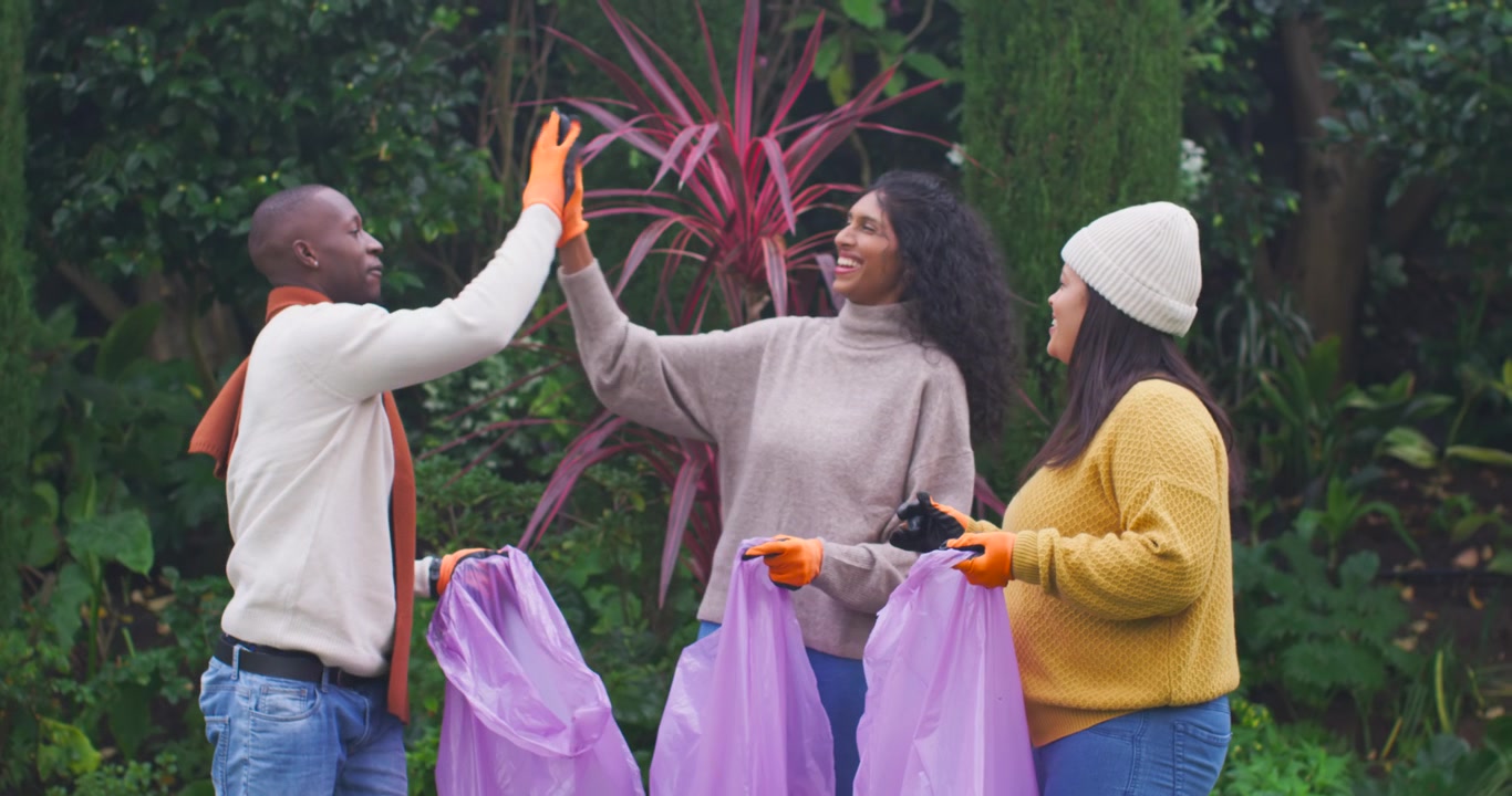 African American friends smiling high-fiving holding purple bags and orange gloves in park to clean