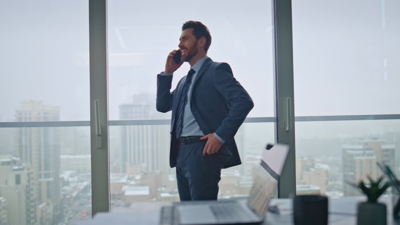 gerente experimentado hablando teléfono móvil en el gabinete de grandes ventanas. llamada de hombre feliz