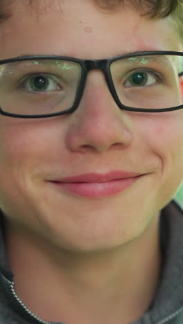 primer plano de un niño con labios rosados, con gafas y una camiseta gris, mirando directamente a la cámara con una ligera sonrisa, está rodeado de vegetación borrosa