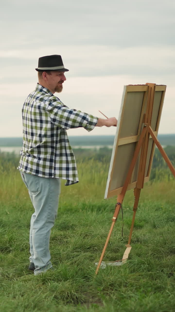 검은 모자, 체크색 셔츠, 청바지를 입은 장인 한 명이 평화로운 잔디 한가운데에 큰 캔버스를 활기차게 칠하고 있습니다.