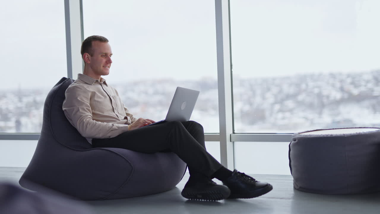 Middle-aged man sitting in chair works on his laptop. Male employee working in comfortable office atmosphere. Side view. Panoramic windows in blur at backdrop.