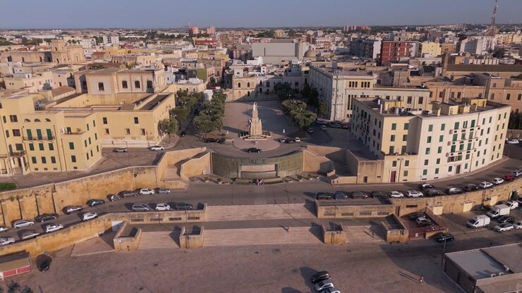 Ripresa aerea in volo verso la Fontana dell'Impero e il Monumento ai Caduti, circondati da architettura storica in Puglia