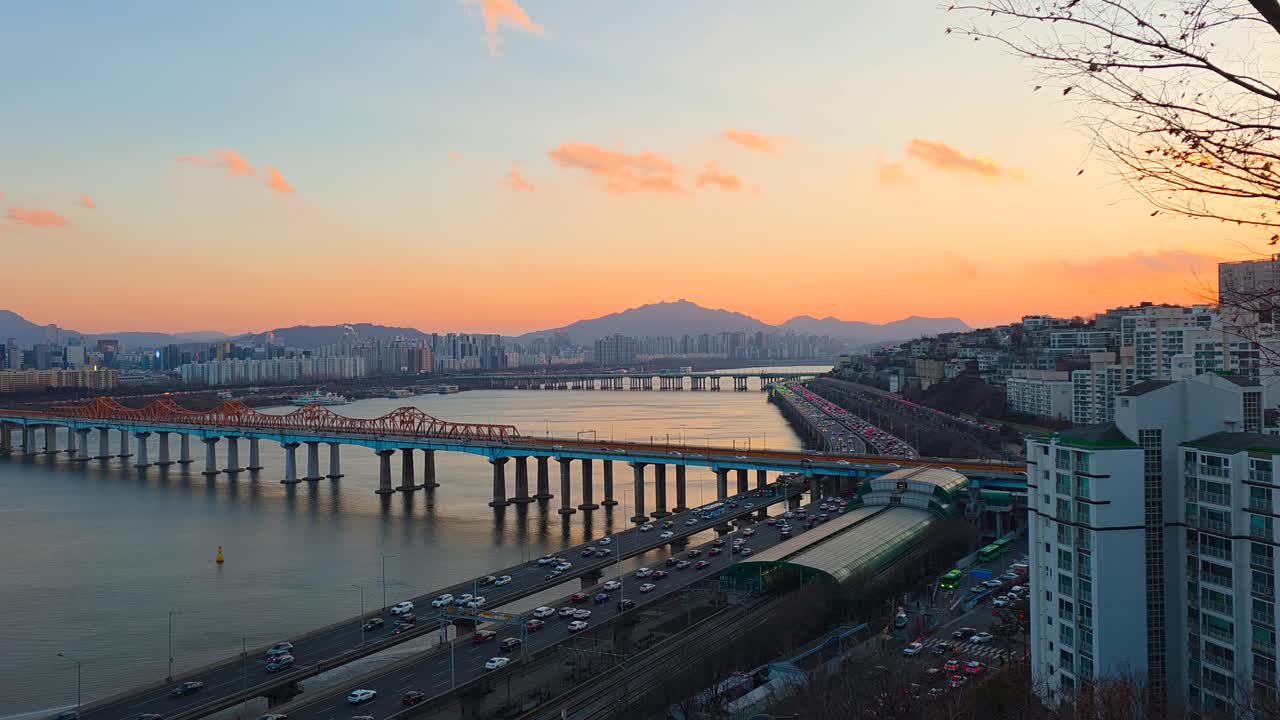 Cielo colorato arancione al tramonto sul fiume Han a Seul, con il Ponte Donho, il traffico dell'autostrada Gangbyeonbuk-ro, il paesaggio urbano e uno sfondo montuoso.