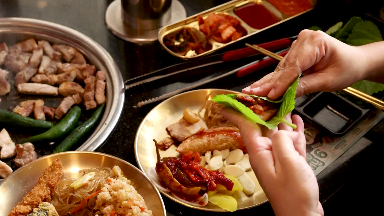 Hands wrap grilled pork with vegetables in leaf at a well-lit Korean barbecue restaurant table