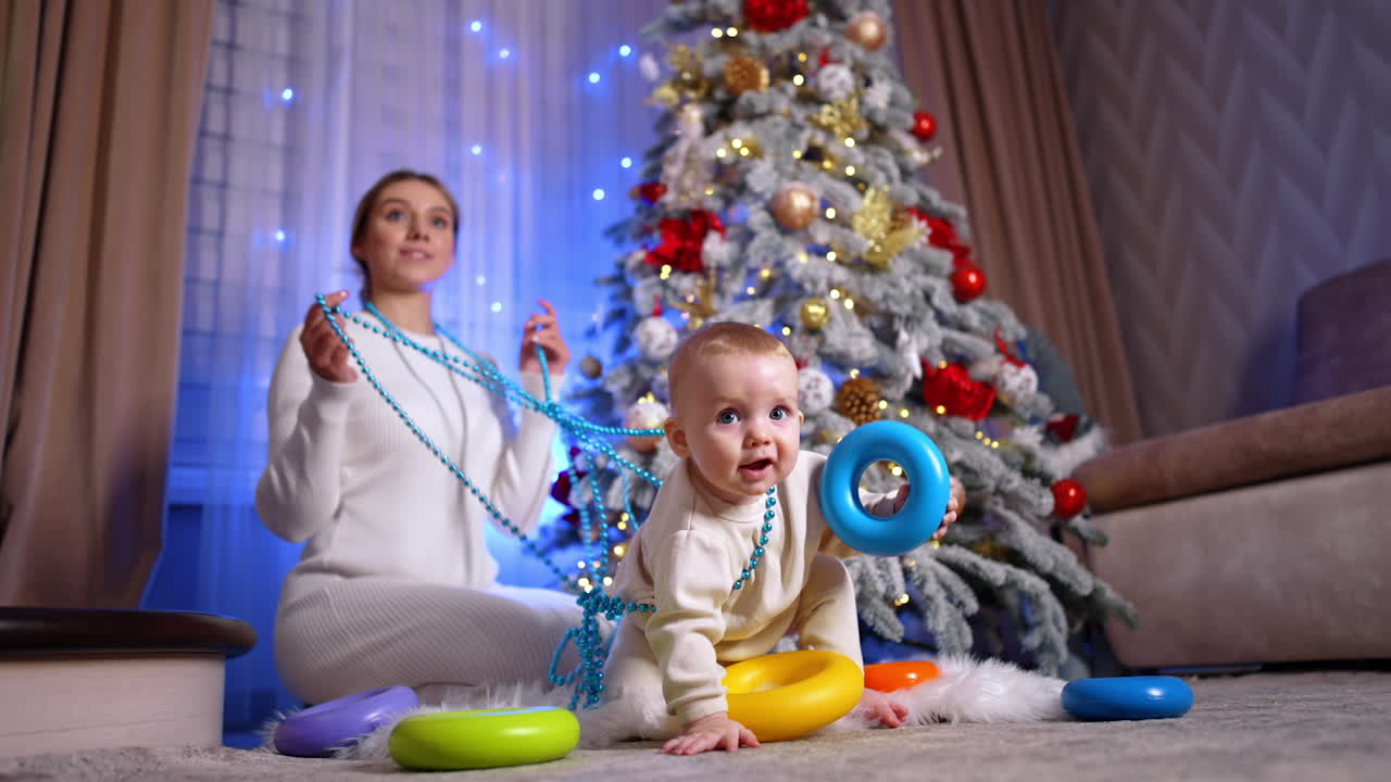 Lovely Caucasian baby is playing with pyramid rings scattered on the floor. Mom sits behind untangling the Christmas beads decoration. Low angle view.