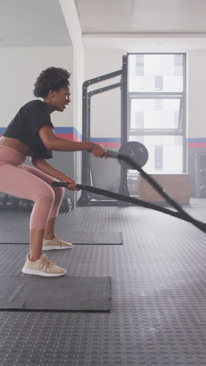Vertical video of determined african american woman working out at a gym with battling ropes