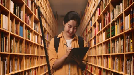estudiante asiática con una mochila sonriendo y tomando nota en la tableta mientras está de pie en la biblioteca
