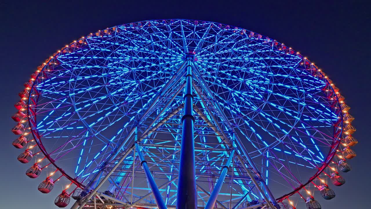 Close-up low angle view of a massive Ferris wheel illuminated with vibrant blue and red LED lights against a dark sky