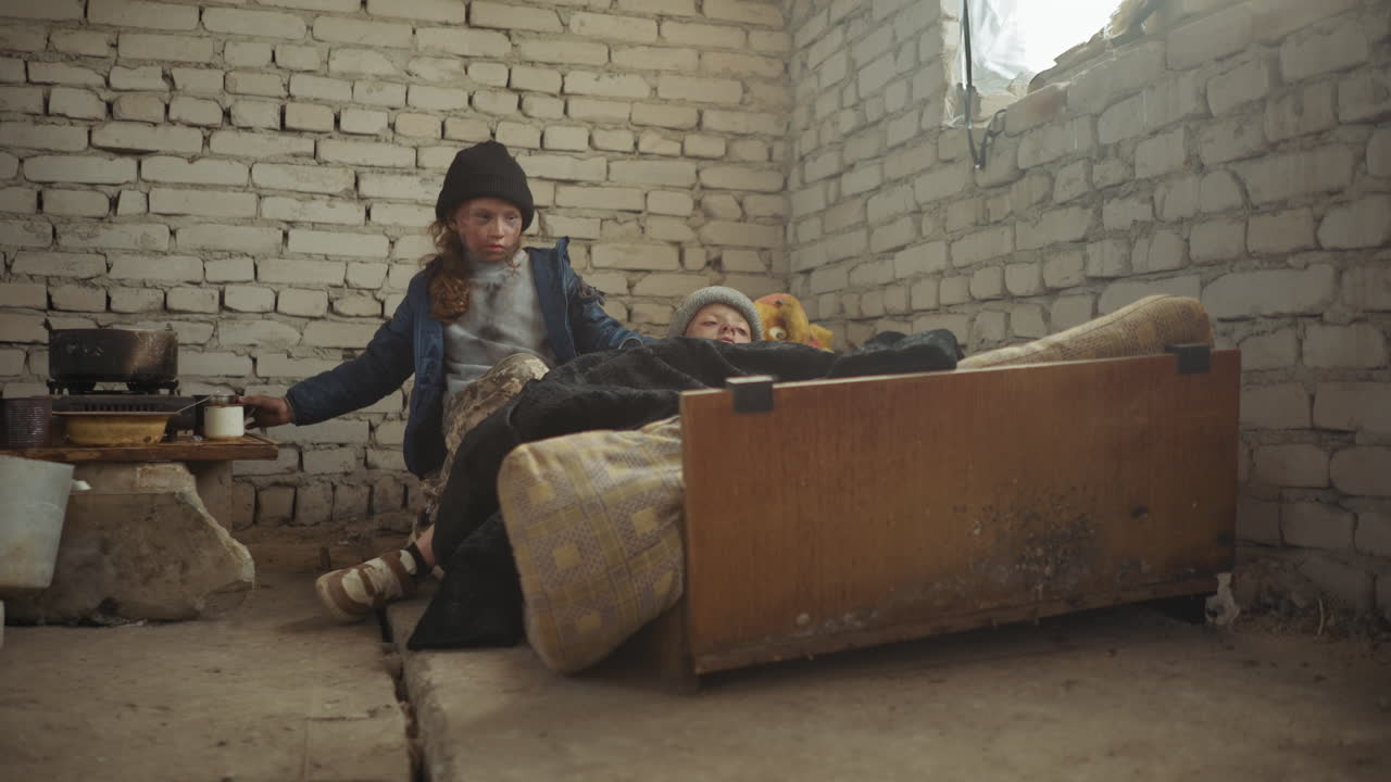 Una niña desplazada, vestida con ropa de invierno, da agua a su hermana enferma sobre un colchón desgastado dentro de un refugio frío de ladrillo; luego, coloca una taza de metal en la mesa cerca de ollas y una estufa, demostrando cuidado y supervivencia.