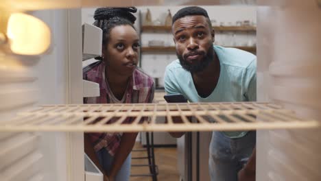 african young couple looking into empty refrigerator and ordering food delivery on smartphone