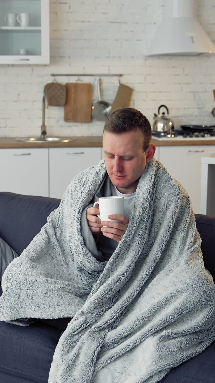 Ill man drinking hot beverage at home. Unwell young man covered with warm blanket sitting on a sofa in the kitchen. Coronavirus pandemic. Vertical video