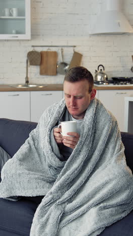 ill man drinking hot beverage at home. unwell young man covered with warm blanket sitting on a sofa in the kitchen. coronavirus pandemic. vertical video