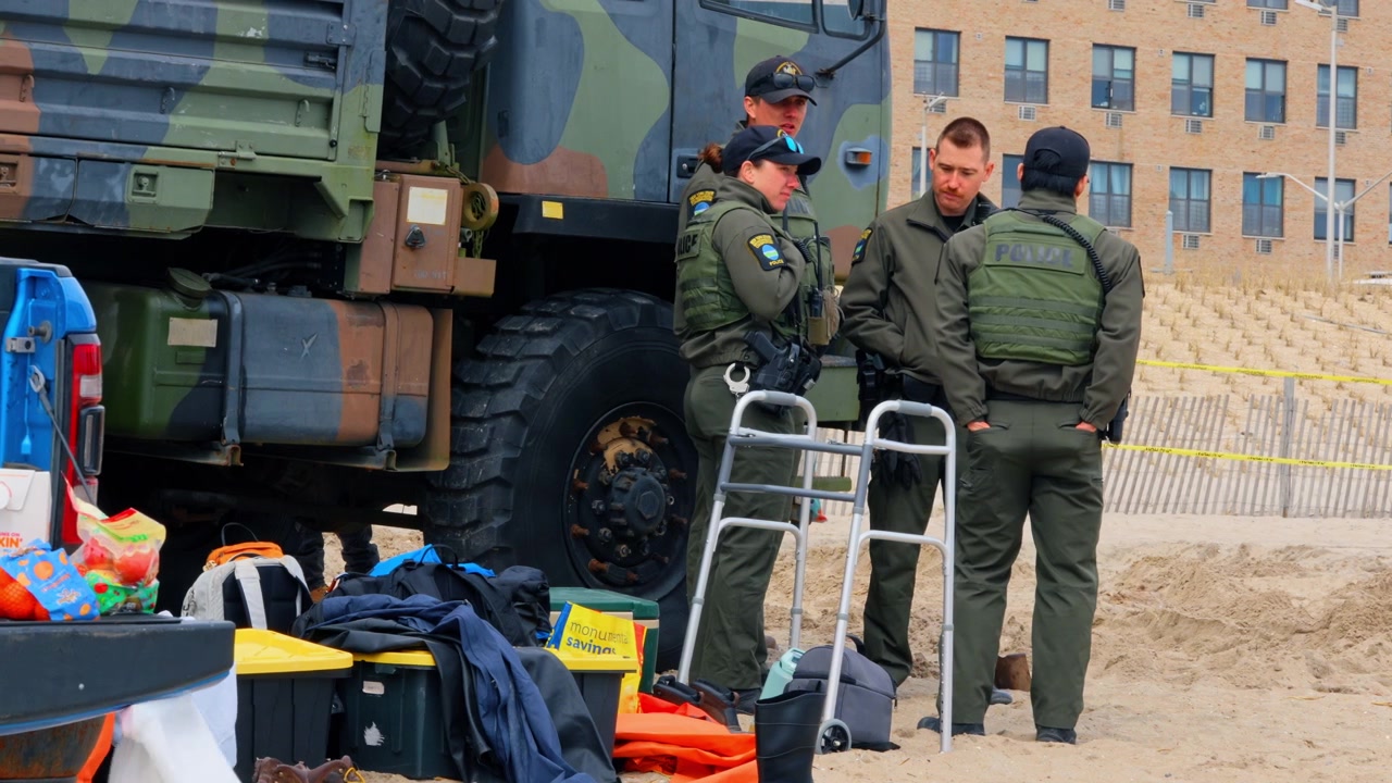 New York, ABD, 1 Nisan 2026: Polis memurları kumlu plajda balina karaya vurma alanını izliyor. Kolluk kuvvetleri personeli, bir deniz memelisi soruşturması sırasında askeri bir kamyonun yanında zeytin yeşili üniformalarıyla duruyor.