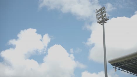 artsy shot of a light pole or tower at a sports stadium, with the corner of the roof of the stand below and white fluffy clouds passing by