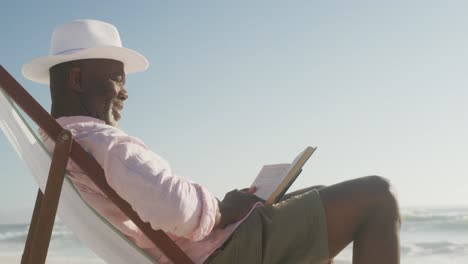 Lächelnder-älterer-Afroamerikanischer-Mann,-Der-Auf-Der-Sonnenbank-Liegt-Und-Am-Sonnigen-Strand-Ein-Buch-Liest