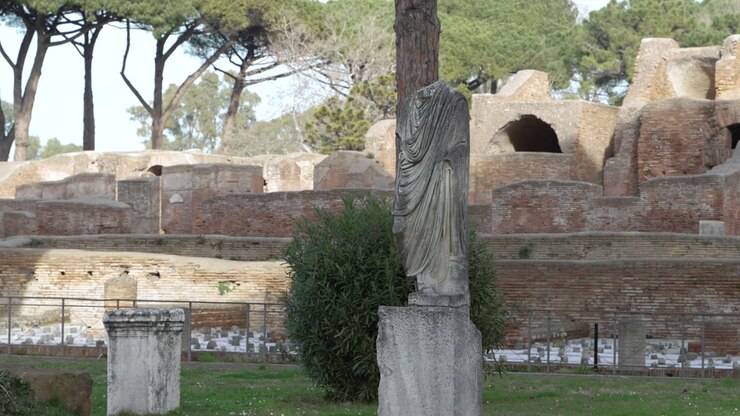Statua drappeggiata senza testa su un piedistallo in una tranquilla area erbosa con portici in mattoni e pini domestici sullo sfondo