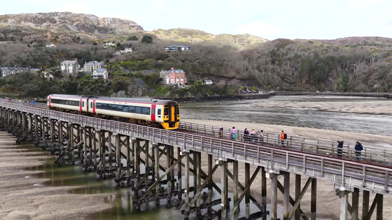 Поезд Transport for Wales пересекает культовый мост Бармут над эстуарием Мауддах, снято с дрона, с обширными прибрежными видами, драматическими пейзажами и потрясающими образцами валлийского железнодорожного строительства.