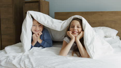 cute funny little sister and brother smiling and looking out of the blanket while lying on the bed in the morning